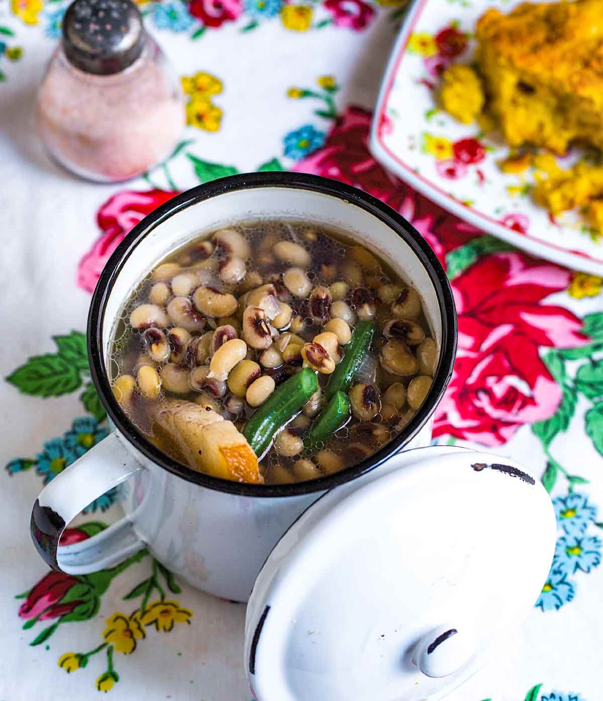 A floral tablecloth with a cellar of pink salt, a plate of cornbread, and a pot of field peas and snap beans along with pot liquor and a chunk of salt pork.