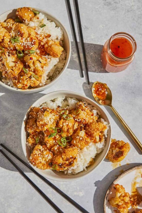 2 white bowls of General-Tso style cauliflower bowls with chopsticks, a jar of extra sauce with spoon and a plate of cauliflower.