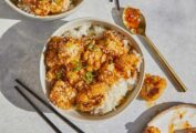 2 white bowls of General-Tso style cauliflower bowls with chopsticks, sauce on a spoon and a plate of cauliflower.