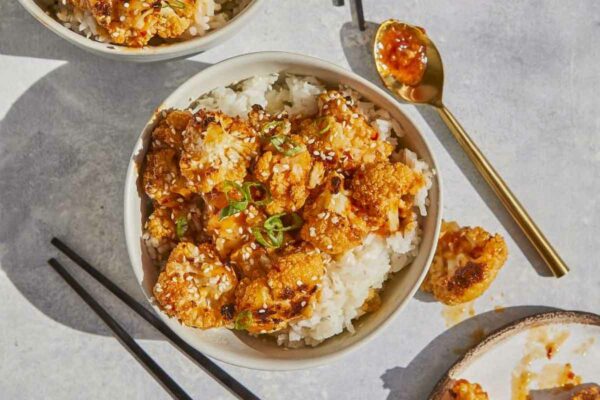 2 white bowls of General-Tso style cauliflower bowls with chopsticks, sauce on a spoon and a plate of cauliflower.