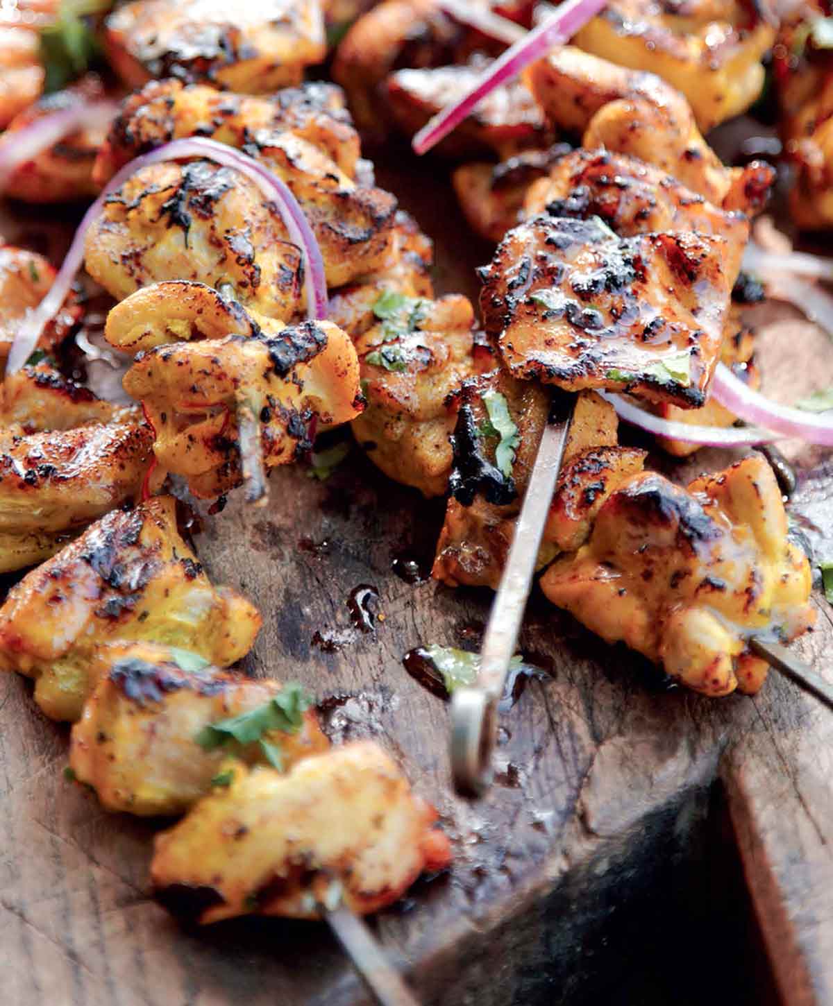 6 grilled chicken skewers piled on a wooden cutting board and sprinkled with slices of red onion and cilantro.