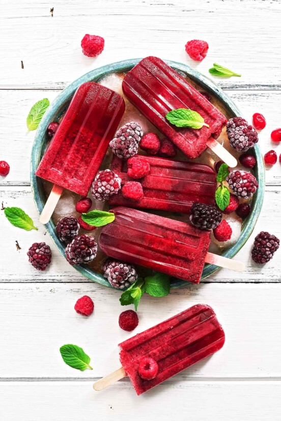 White barn board with a green bowl filled with ice and frozen berries, and 5 deep red raspberry hard seltzer popsicles lying on top, garnished with fresh mint leaves.