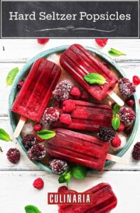 White barn board with a green bowl filled with ice and frozen berries, and 5 deep red raspberry hard seltzer popsicles lying on top, garnished with fresh mint leaves.