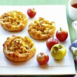 Three honey-ginger apple tarts on a white platter with 4 apples on the side.