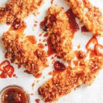 5 panko breaded chicken breasts drizzled with deep red hot sauce and chilli slices, accompanied by a bowl of spiced honey and a honey spoon.