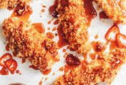 5 panko breaded chicken tenders drizzled with deep red hot sauce and chile slices, with a bowl of more spicy honey and a honey dipper.