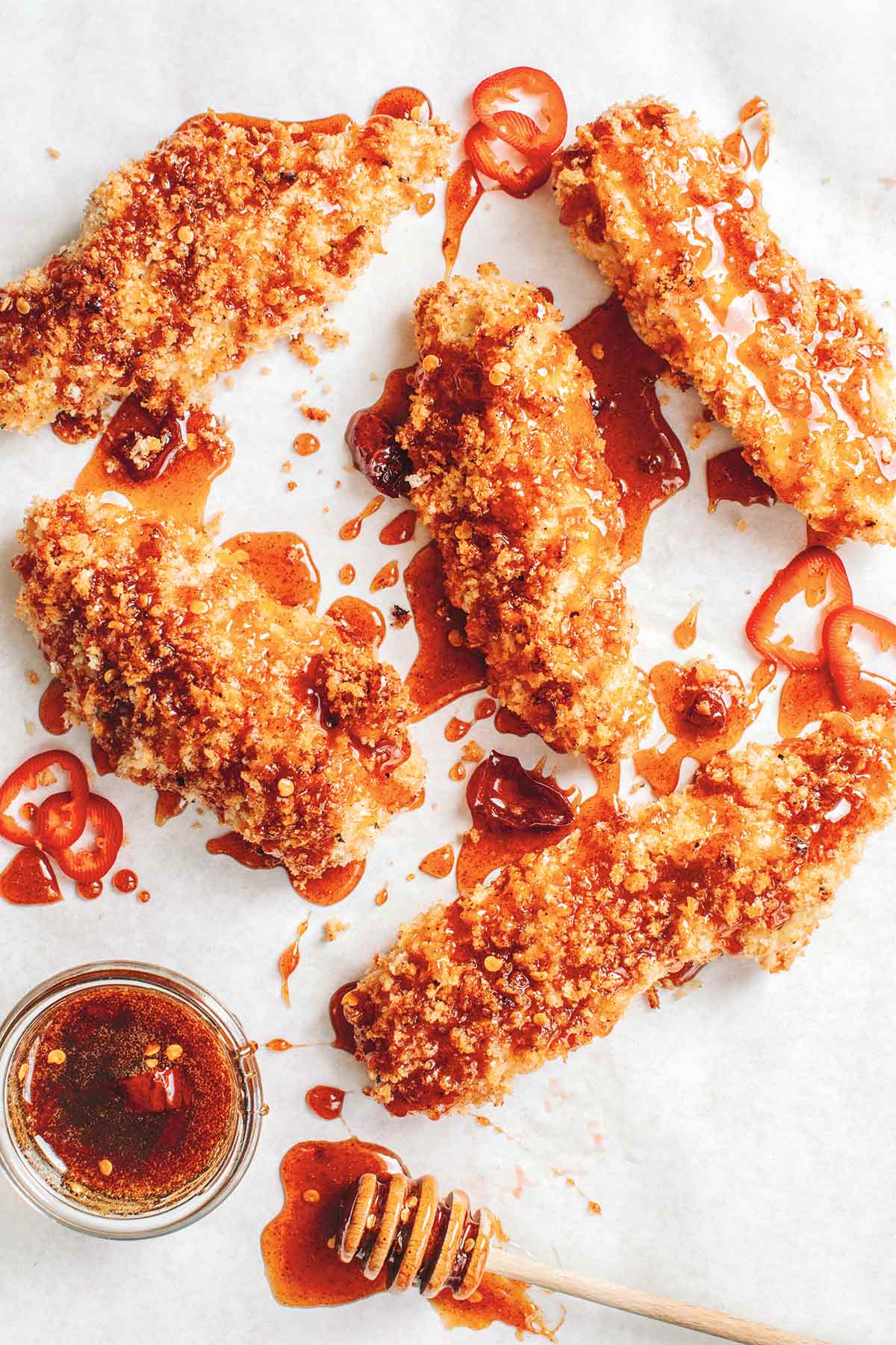 5 panko breaded chicken tenders drizzled with deep red hot sauce and chile slices, with a bowl of more spicy honey and a honey dipper.