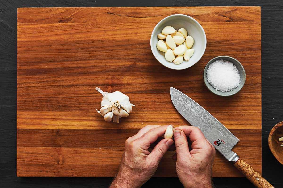 A person peeling a clove of garlic over a cutting board with a knife, garlic, and salt on it.
