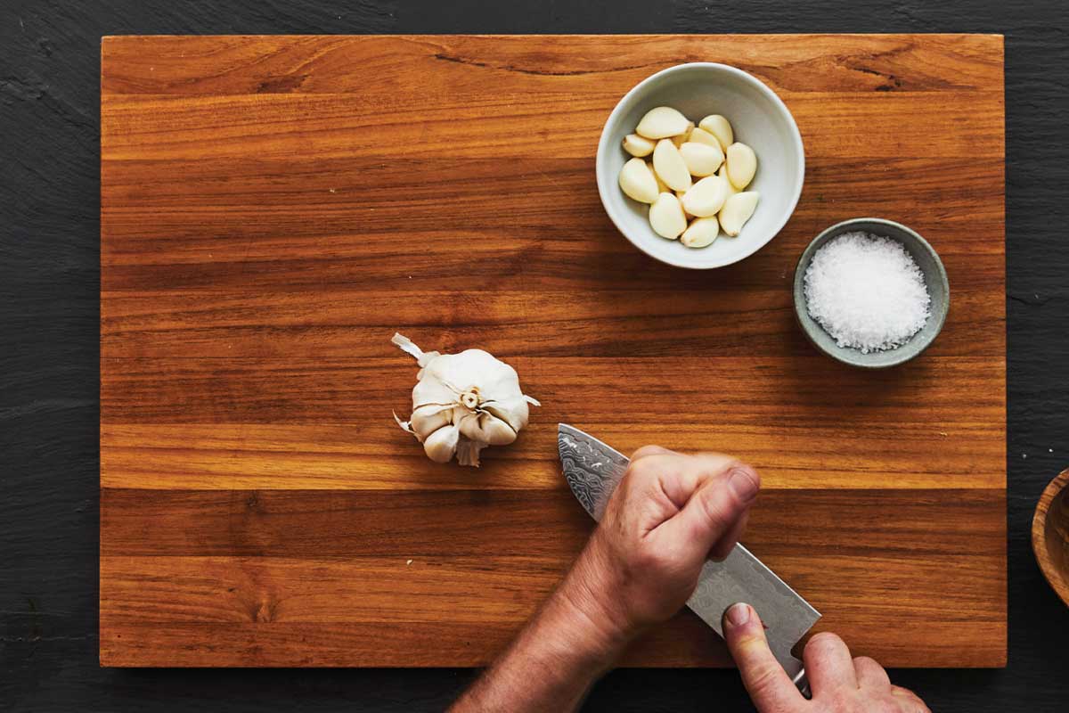 A person smashing a clove of garlic on a cutting board.