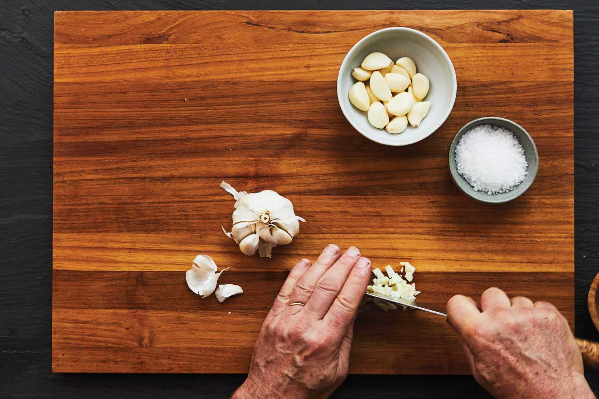 A person mincing a clove of garlic on a cutting board with garlic and a bowl of salt on it.