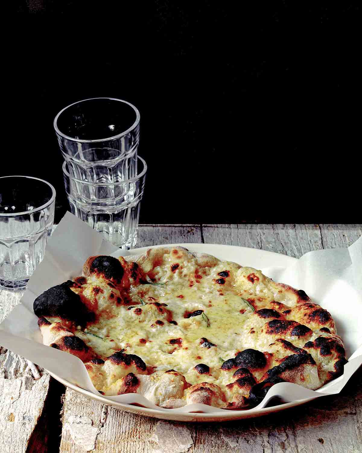 Ett träbord med en vit platta med bakplåtspapper och Jim Laheys vita pizza, med en bunt glas i bakgrunden.