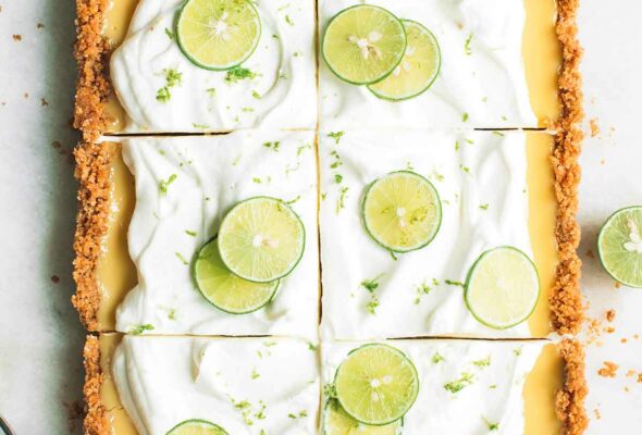 A large rectangular key lime pie cut into sixths, covered with whipping cream and slices of lime, covered with lime zest and sliced limes next to it.