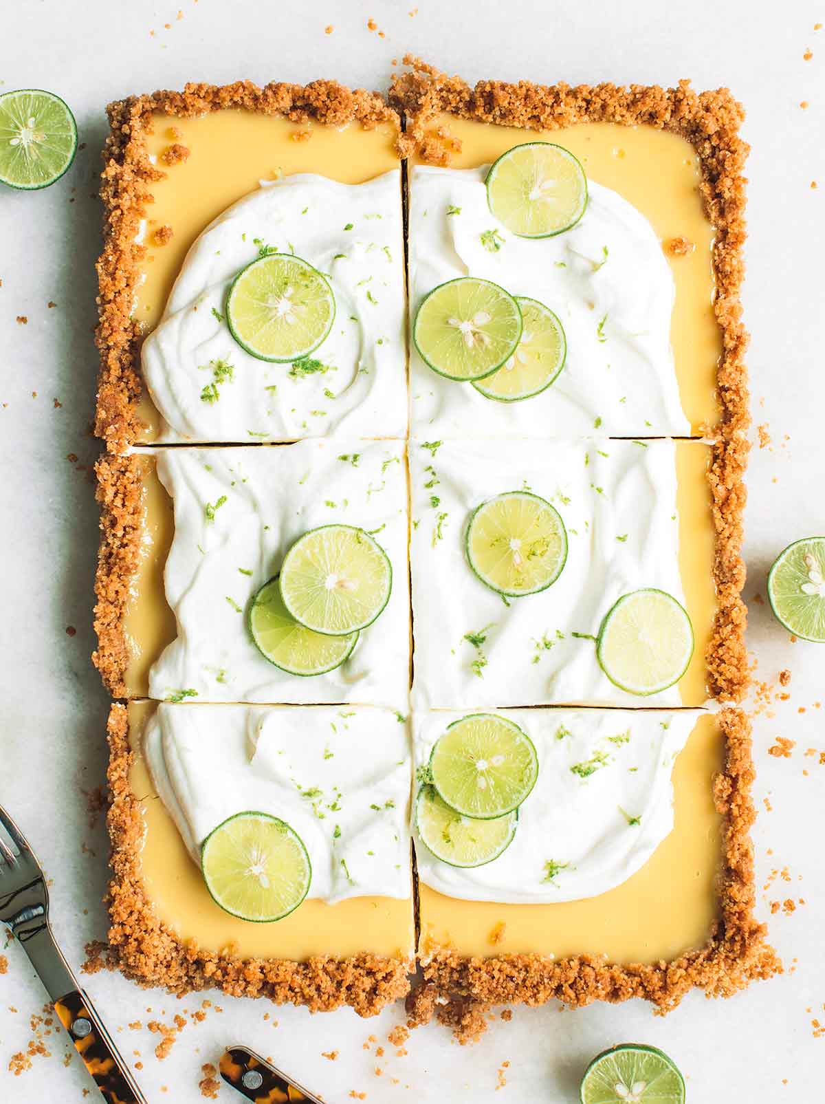 A large rectangular key lime pie cut into sixths, covered with whipping cream and slices of lime, covered with lime zest and sliced limes next to it.