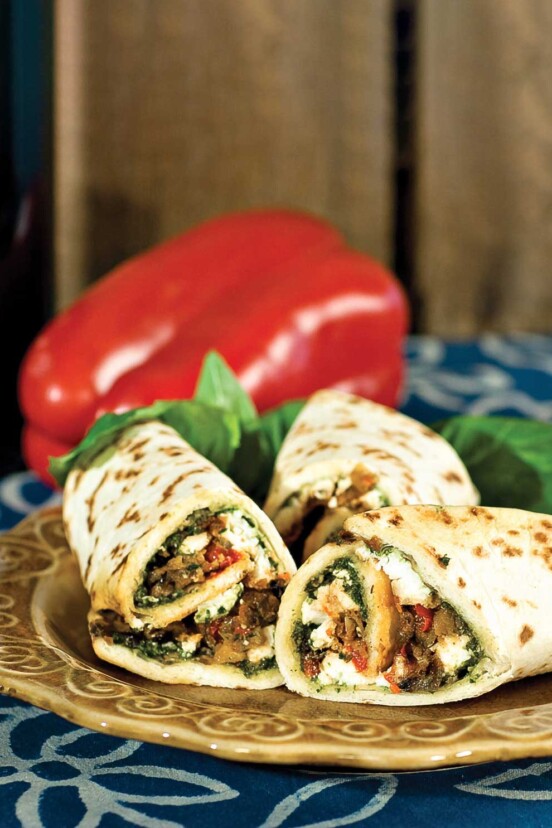 A plate with three eggplant wraps and loose basil leaves, with a whole red pepper in the background