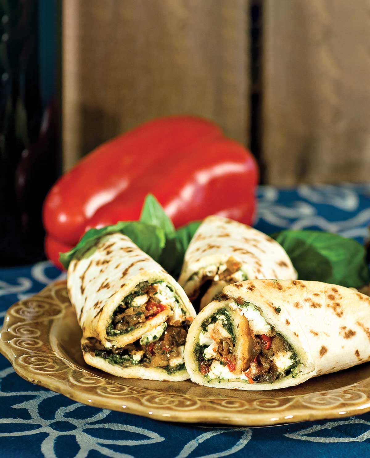 A plate with three eggplant wraps and loose basil leaves, with a whole red pepper in the background