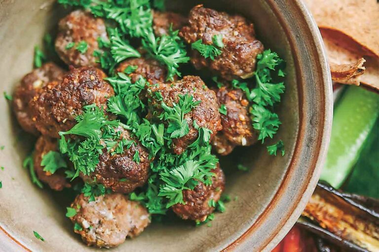 A pottery bowl filled with merguez meatballs, garnished with parsley, surrounded by pitas and roasted red peppers.
