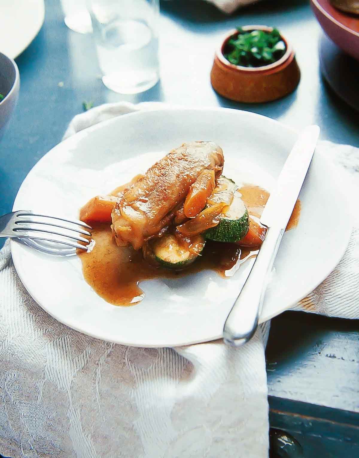En vit tallrik på ett bord med kniv och gaffel, med en portion marockansk bräserad kyckling med dadlar och grönsaker, inklusive morötter och zucchini.