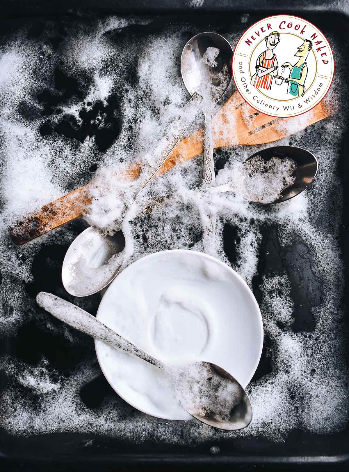 4 silver spoons, a wooden spatula, and a white saucer all covered with suds.