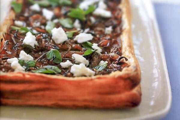 A white rectangular platter with a puff pastry tart filled with caramelized onions, feta cheese, and oregano.