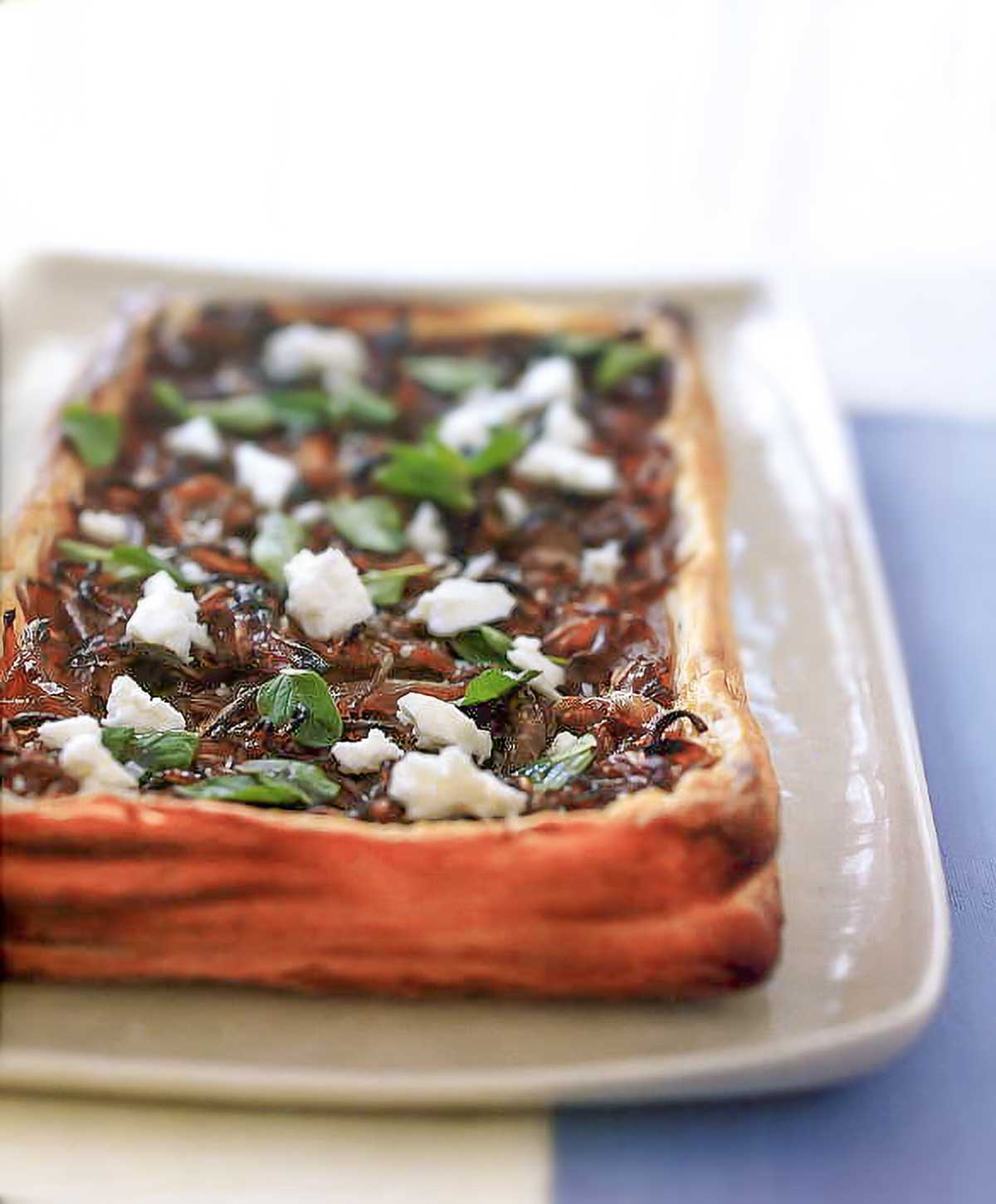 A white rectangular platter with a puff pastry tart filled with caramelized onions, feta cheese, and oregano.