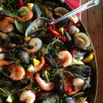 A large Dutch oven full of shrimp, mussels, red peppers, peas, lemon slices, parsley, bay leaves, and onion with a serving spoon.