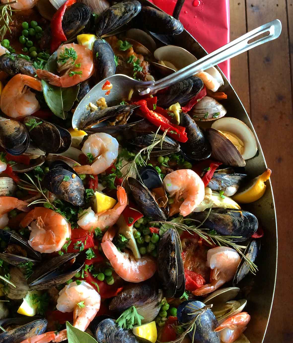 A large Dutch oven full of shrimp, mussels, red peppers, peas, lemon slices, parsley, bay leaves, and onion with a serving spoon.
