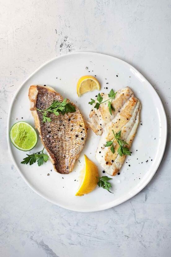 A white plate white 2 pieces of fish, one sliced and one whole with crisp skin attached. Garnished with slices of lemon, lime, and parsley.