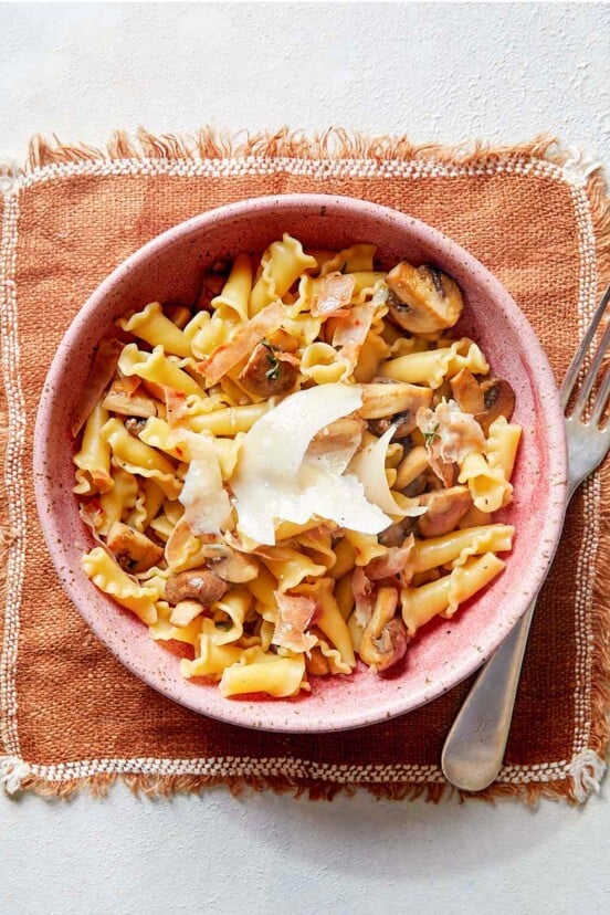 A pink bowl of short pasta with mushrooms, prosciutto, and Parmesan cheese on a placemat with a fork.