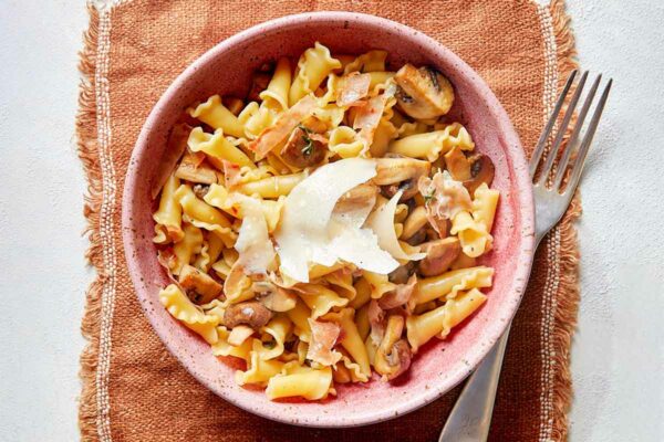 A pink bowl of short pasta with mushrooms, prosciutto, and Parmesan cheese on a placemat with a fork.