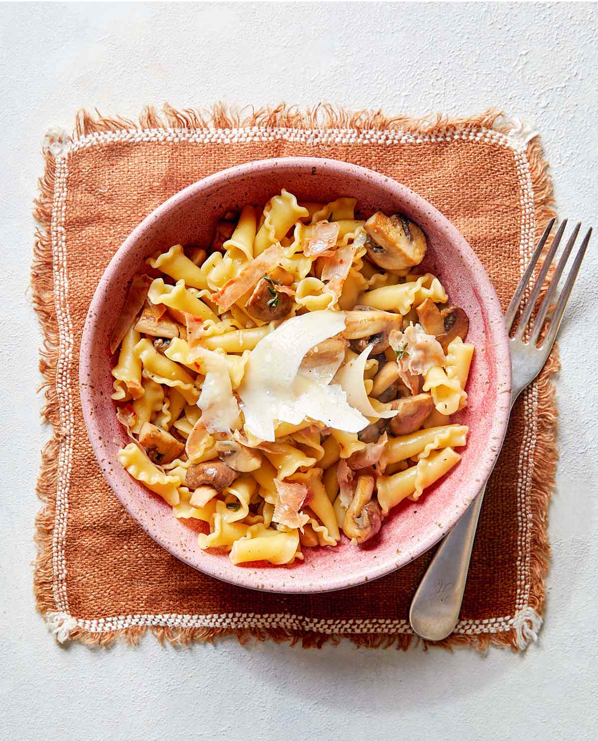 A pink bowl of short pasta with mushrooms, prosciutto, and Parmesan cheese on a placemat with a fork.