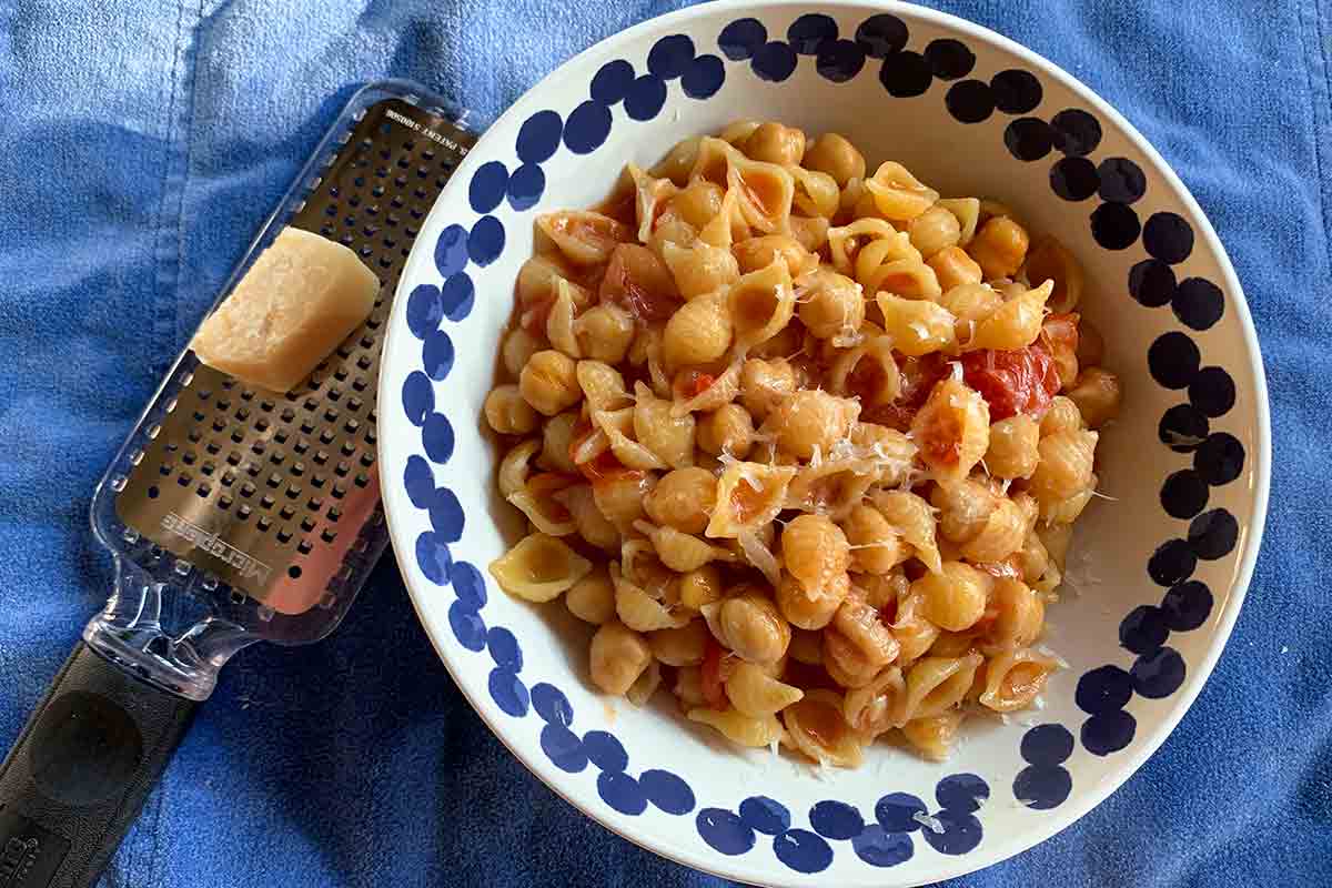 A blue and white bowl filled with pasta shells, tomatoes, chickpeas, and shavings of Parmesan, beside a grater and a chunk of Parmesan.