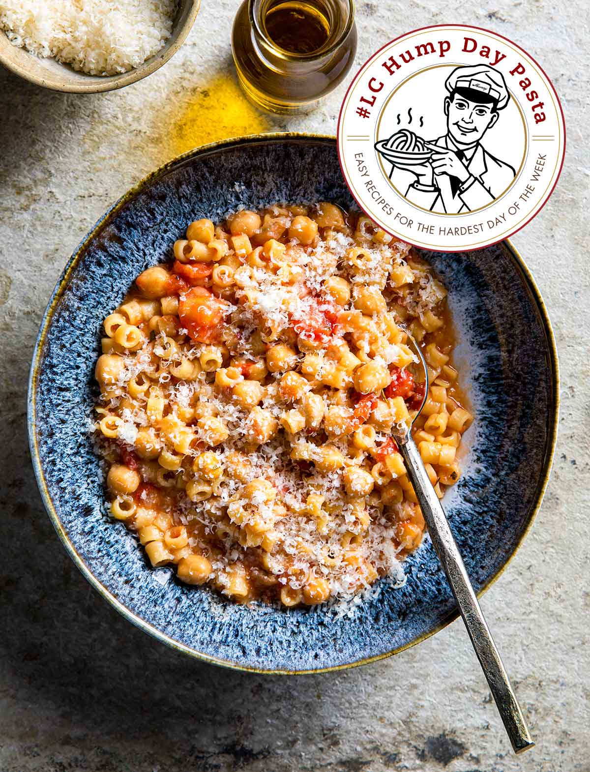 A blue bowl filled with chickpeas, diced tomatoes, ditalini pasta, and parmesan, flanked with a crueset of olive oil and a bowl of Parmesan.