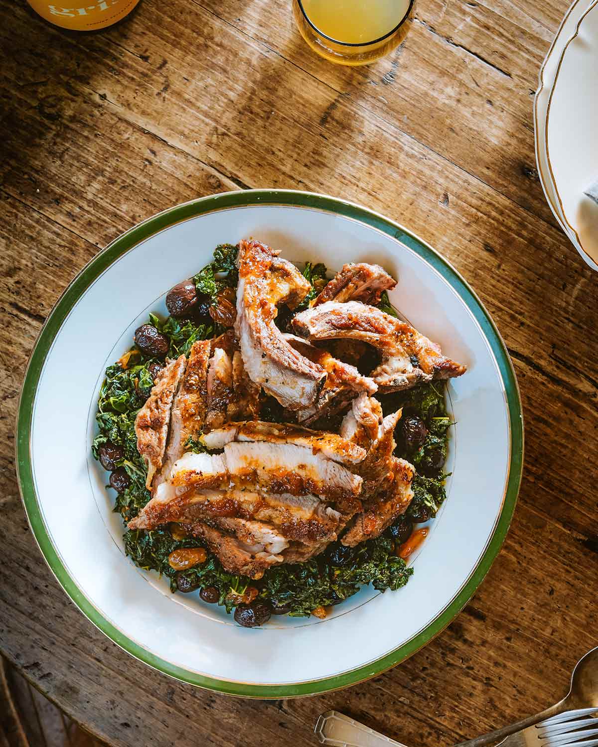 A green and white platter with wilted kale, raisins, olives, and a pile of sliced pork.