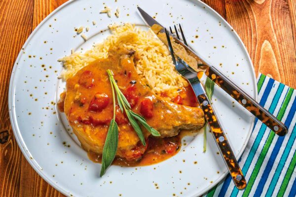A plate with a pork chop and tomato-sage gravy and rice, garnished with sage. Flanked by a knife and fork.