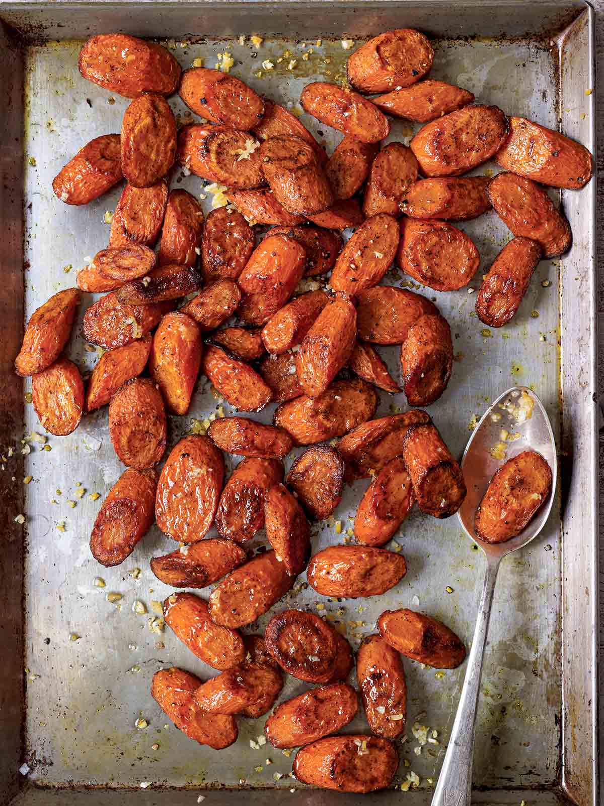 A large metal sheet pan filled with slices of roasted carrots with allspice, sea salt and preserved lemons, with a serving spoon.