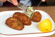A close up of an oval plate with 3 pastéis de bacalhau, or Portuguese salt cod fritters, and a lemon wedge, one is being cut into with a knife and fork.