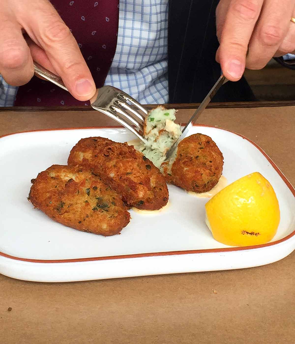 A close up of an oval plate with 3 pastéis de bacalhau, or Portuguese salt cod fritters, and a lemon wedge, one is being cut into with a knife and fork.