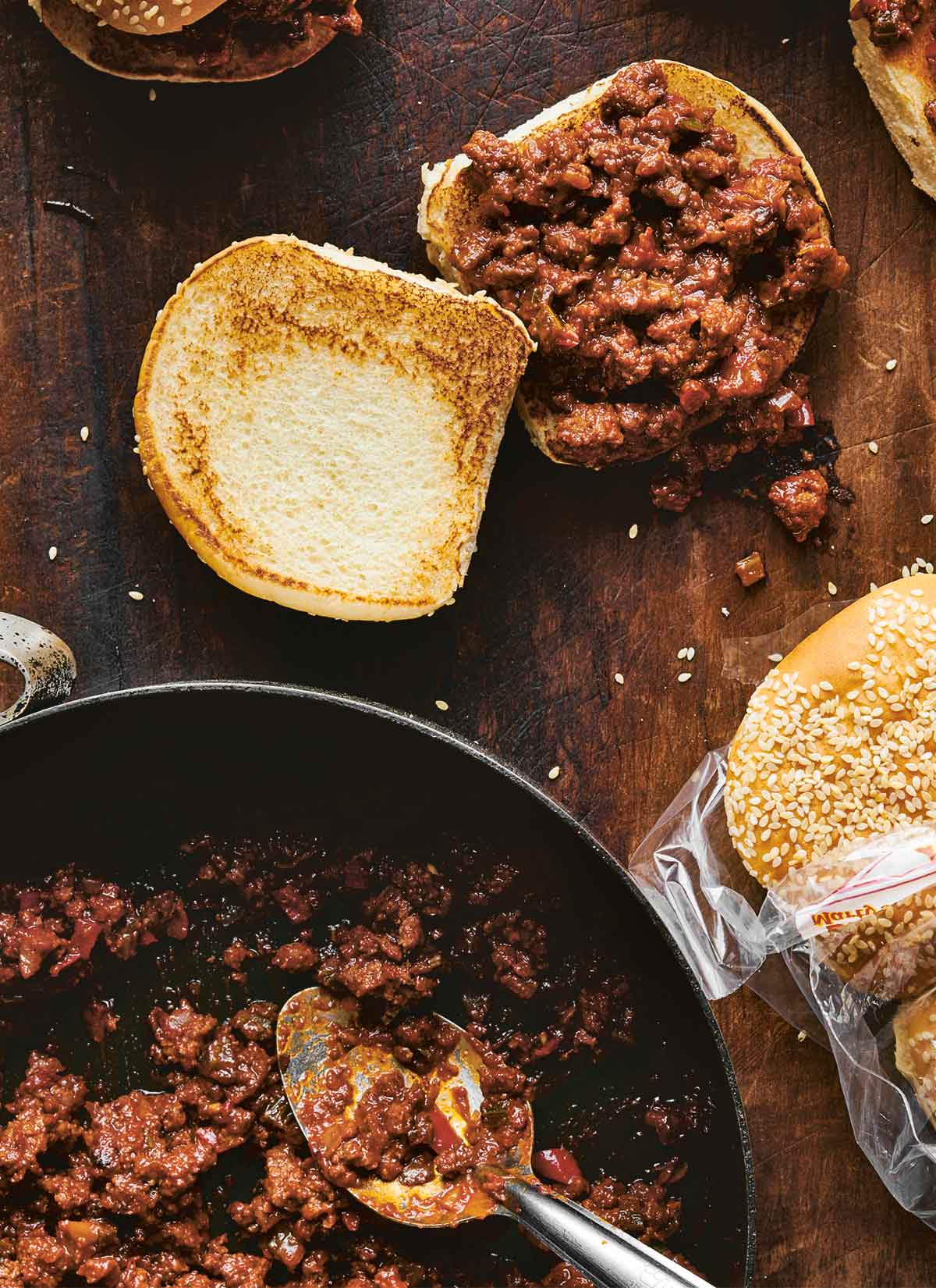 A skillet filled with sloppy Joe meat mixture, flanked by a filled, untapped sandwich with a toasted top and a bag of sesame seed buns.