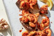 A large white serving tray with 3 pieces of toasted bread covered with dark red sauced shrimp, a lemon slice and a drizzle of sauce. A large basket of garlic and garlic cloves sits beside it.