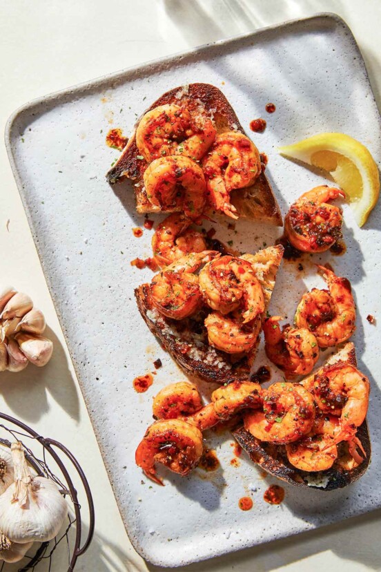 A large white serving tray with 3 pieces of toasted bread covered with dark red sauced shrimp, a lemon slice and a drizzle of sauce. A large basket of garlic and garlic cloves sits beside it.