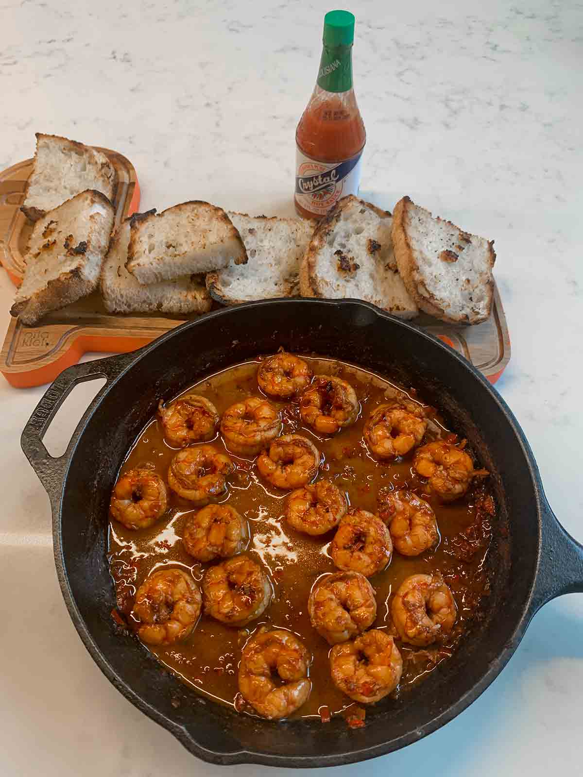 A large cast-iron skillet filled with shrimp in a dark red sauce, beside a cutting board with toasted slices of bread and a bottle of hot sauce.