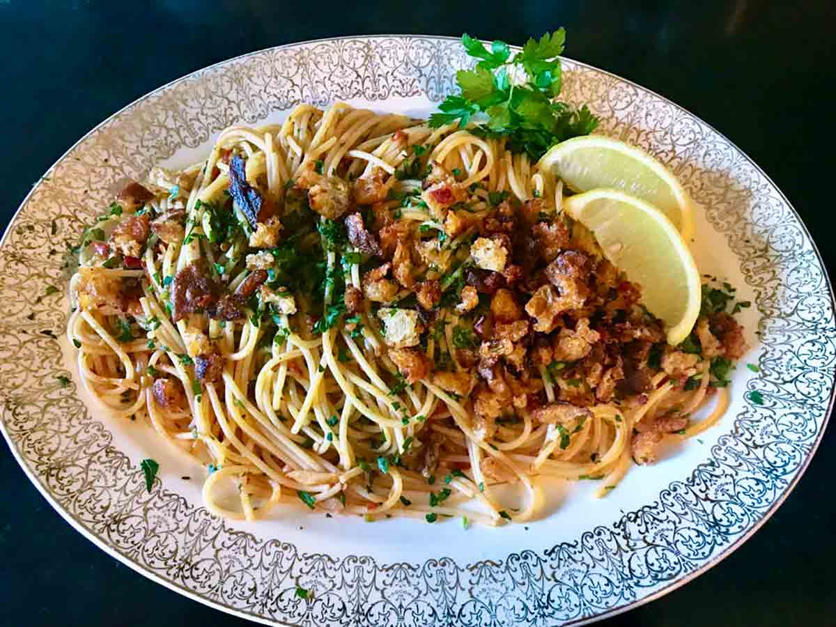 An oval plate filled with Sriracha and lemon spaghetti with Chile pangrattato, topped with crispy bread, parsley, chile flakes, and 2 wedges of lemon on the side.