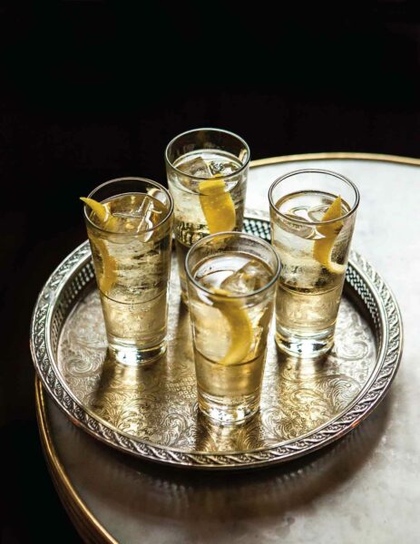4 delicate tumblers filled with St. Germain Cocktail with a little ice and a lemon twist, sitting on a silver tray on a marble table.