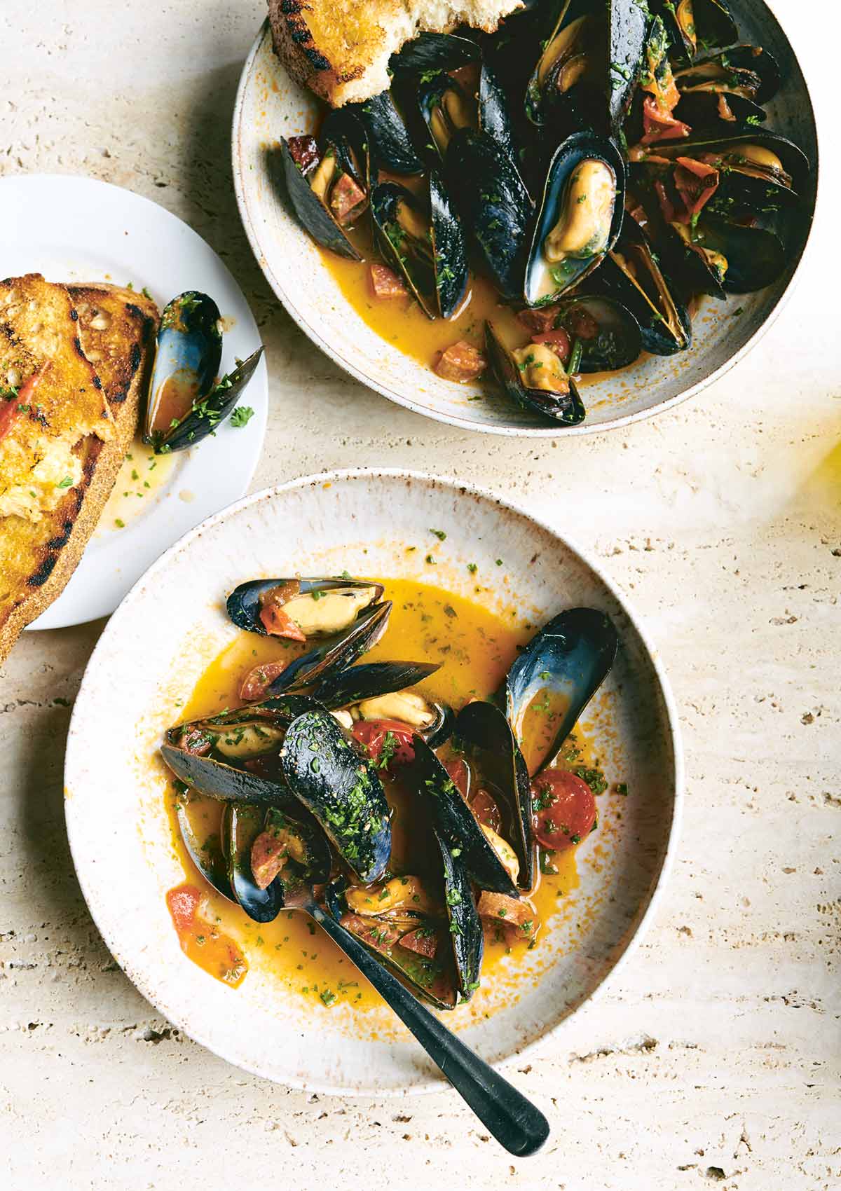 2 white plates with mussels in sauce, with tomatoes, chorizo, and parsley. A piece of buttery toasted bread to the side.