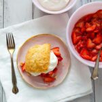 A strawberry shortcake on a pink plate with a bowl of strawberries and a bowl of cream beside it.