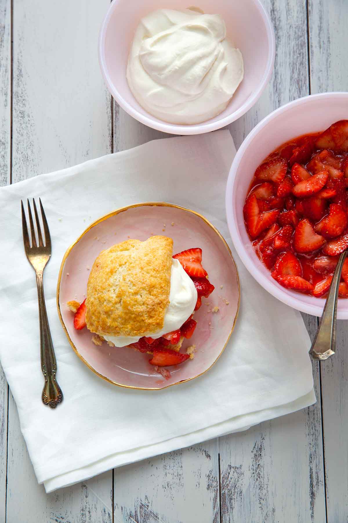 A strawberry shortcake on a pink plate with a bowl of strawberries and a bowl of cream beside it.
