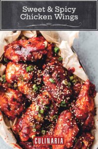 A basket of chicken wings with wax paper, covered with a sticky red sauce, sesame seeds, and green onion slices.