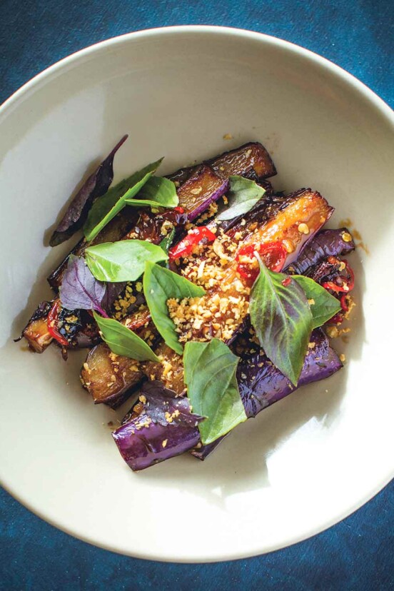 A white bowl filled with slices of eggplant, rings of chiles, covered with chopped garlic and purple basil.