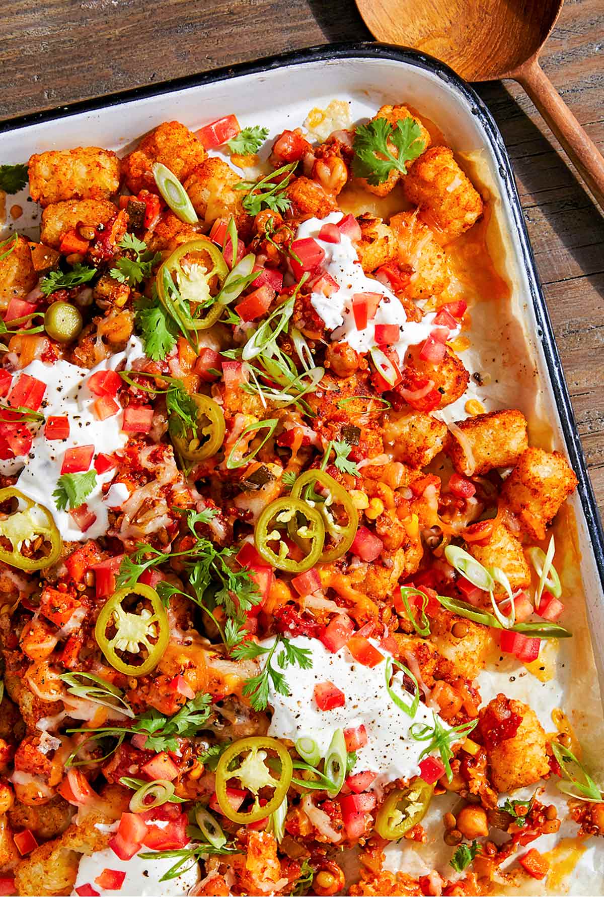 A large rectangle casserole pan, filled with tater tot nachos topped with sour cream, tomatoes, sour cream, and jalapeños. A wooden serving spoon lays beside the dish.