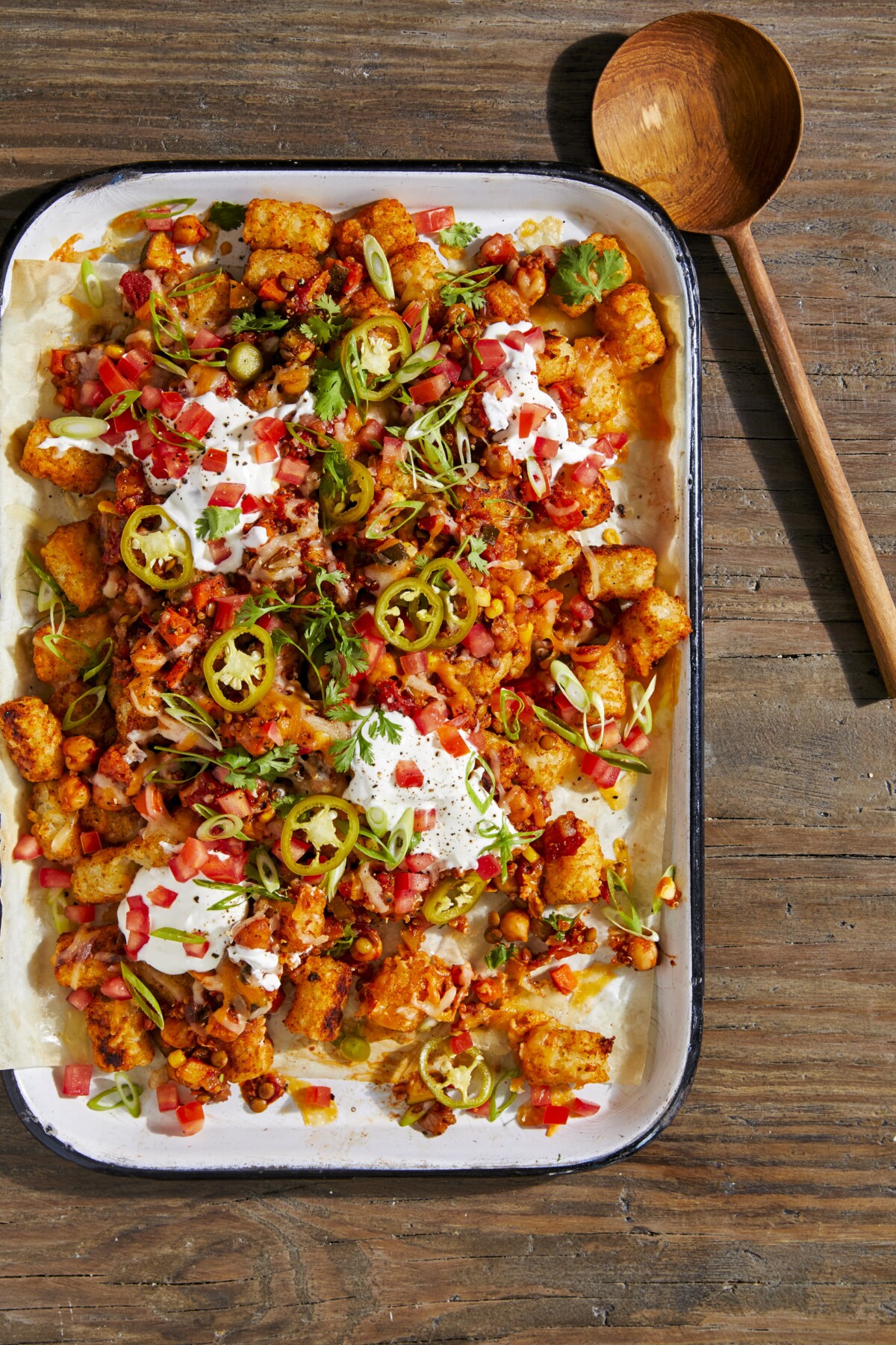 A large rectangle casserole pan, filled with tater tot nachos topped with sour cream, tomatoes, sour cream, and jalapeños. A wooden serving spoon lays beside the dish.